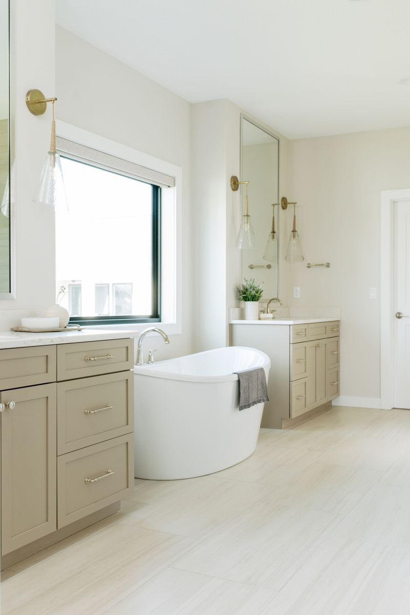 A primary bath in Clive with a freestanding white tub set between greige vanities under a black framed window.