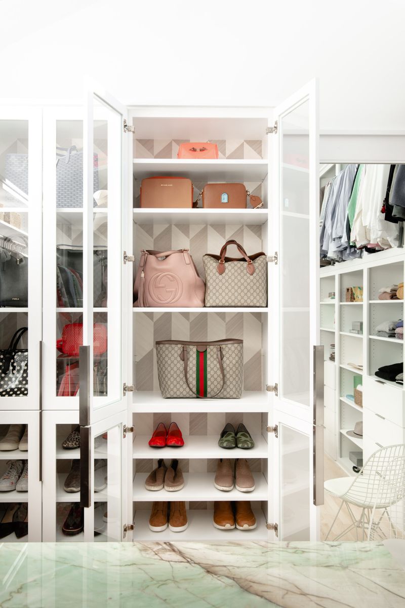 A white walk in closet in Des Moines with glass front cabinets displaying designer handbags backed by geometric tan wallpaper.