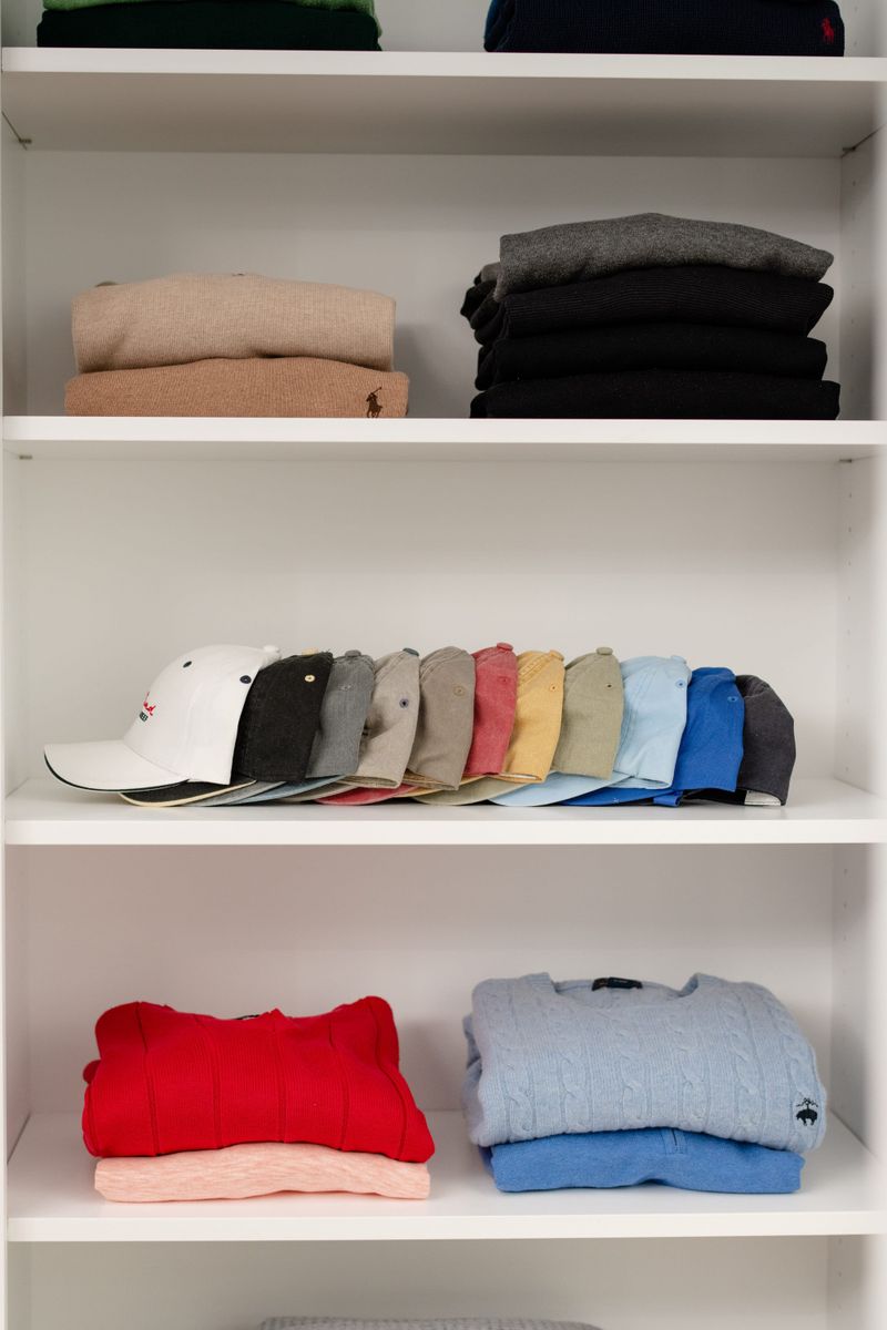 A walk in closet in Des Moines with white adjustable shelves holding a row of ball caps above folded sweaters.