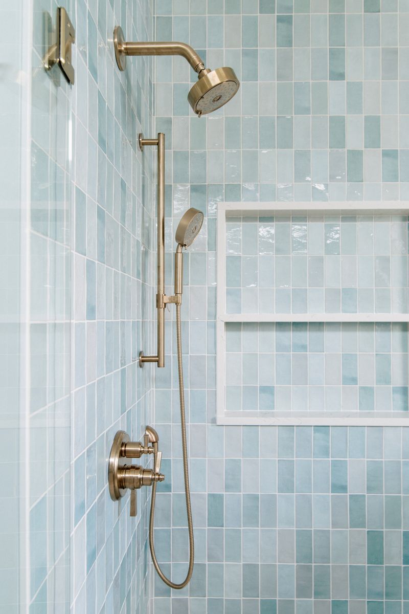 A bathroom shower in Panora with brushed brass fixtures against blue green tile and a stone edged niche.