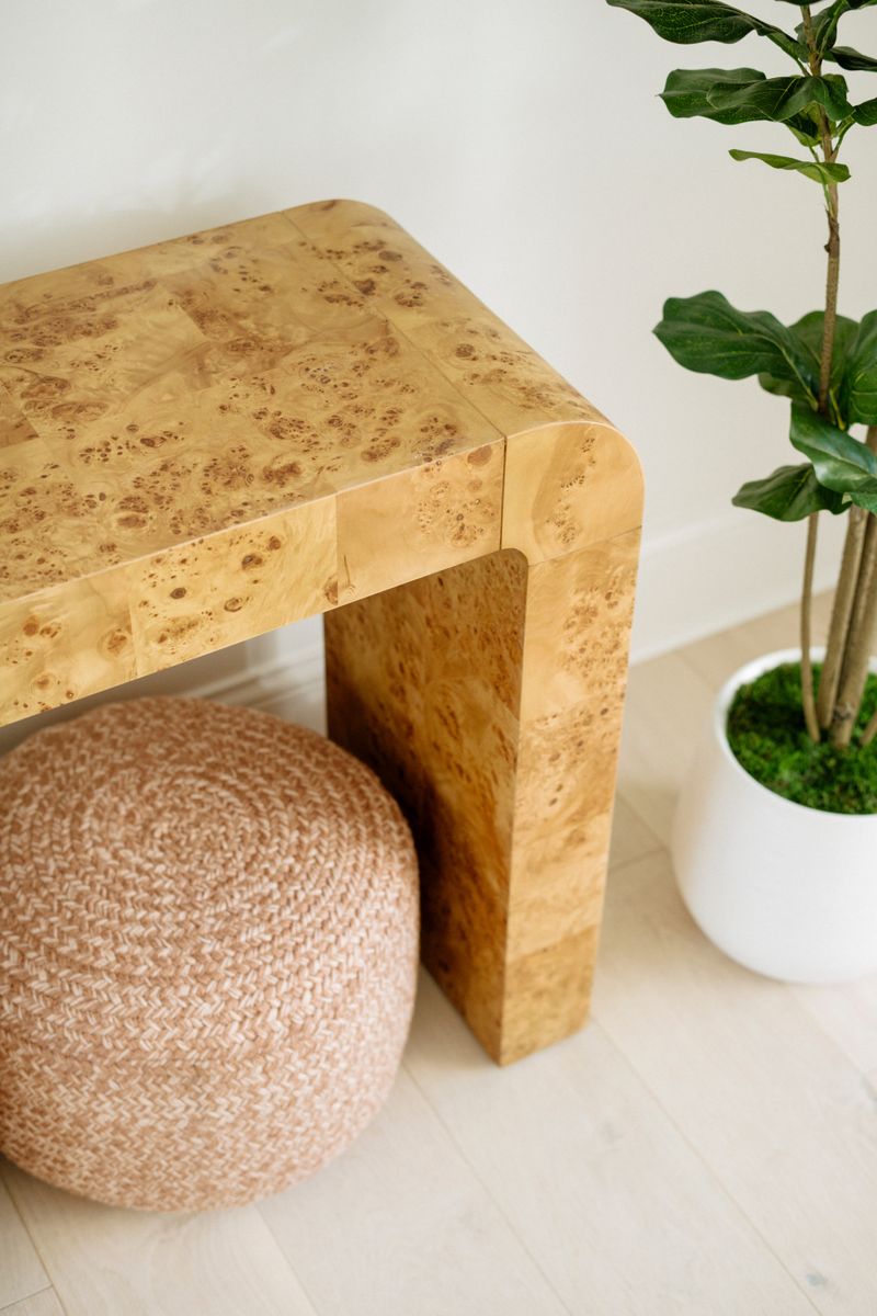 A burl wood console table in a Panora entry, with a woven blush pouf tucked underneath it.