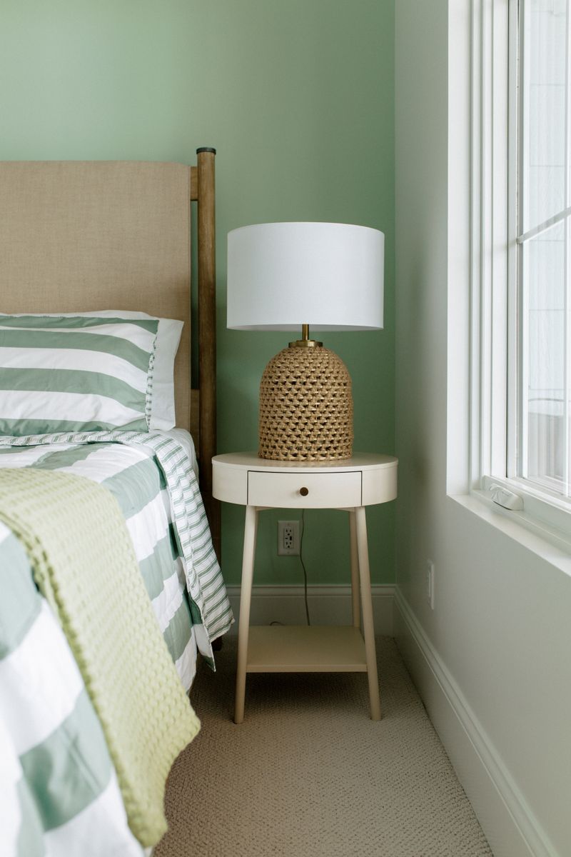 A green bedroom in Panora with a woven rattan lamp on a round cream nightstand.
