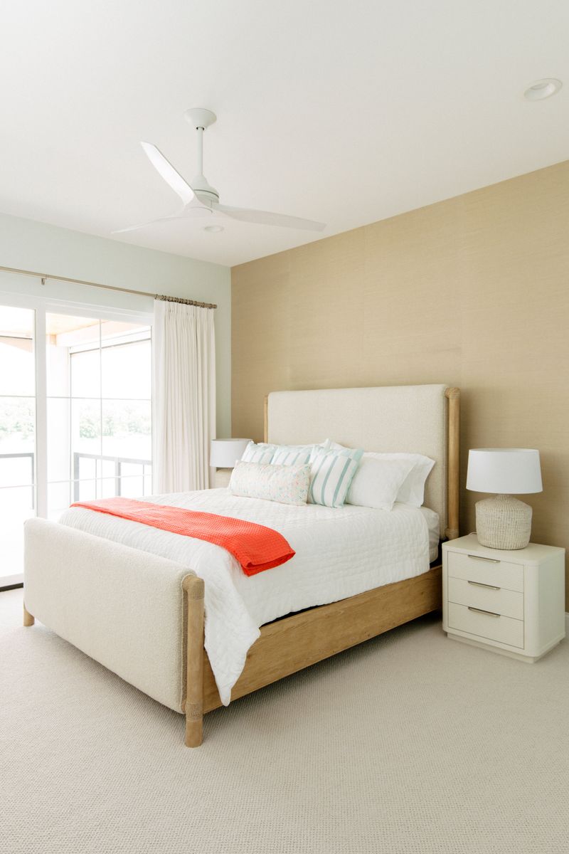 A guest bedroom in Panora with a cream upholstered bed against a grasscloth wall, warmed by a coral throw.