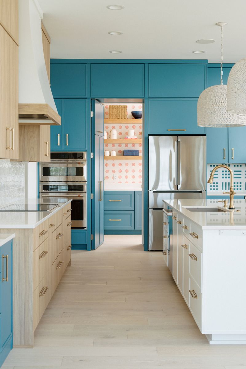 A teal kitchen in Panora with brass hardware and a walk-in pantry papered in pink behind the cabinets.
