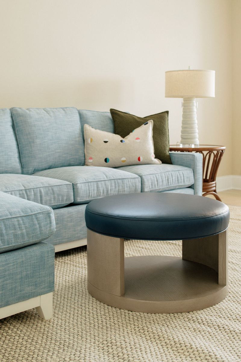 A living room in Panora with a round navy leather ottoman on a pale oak base beside a light blue sectional.