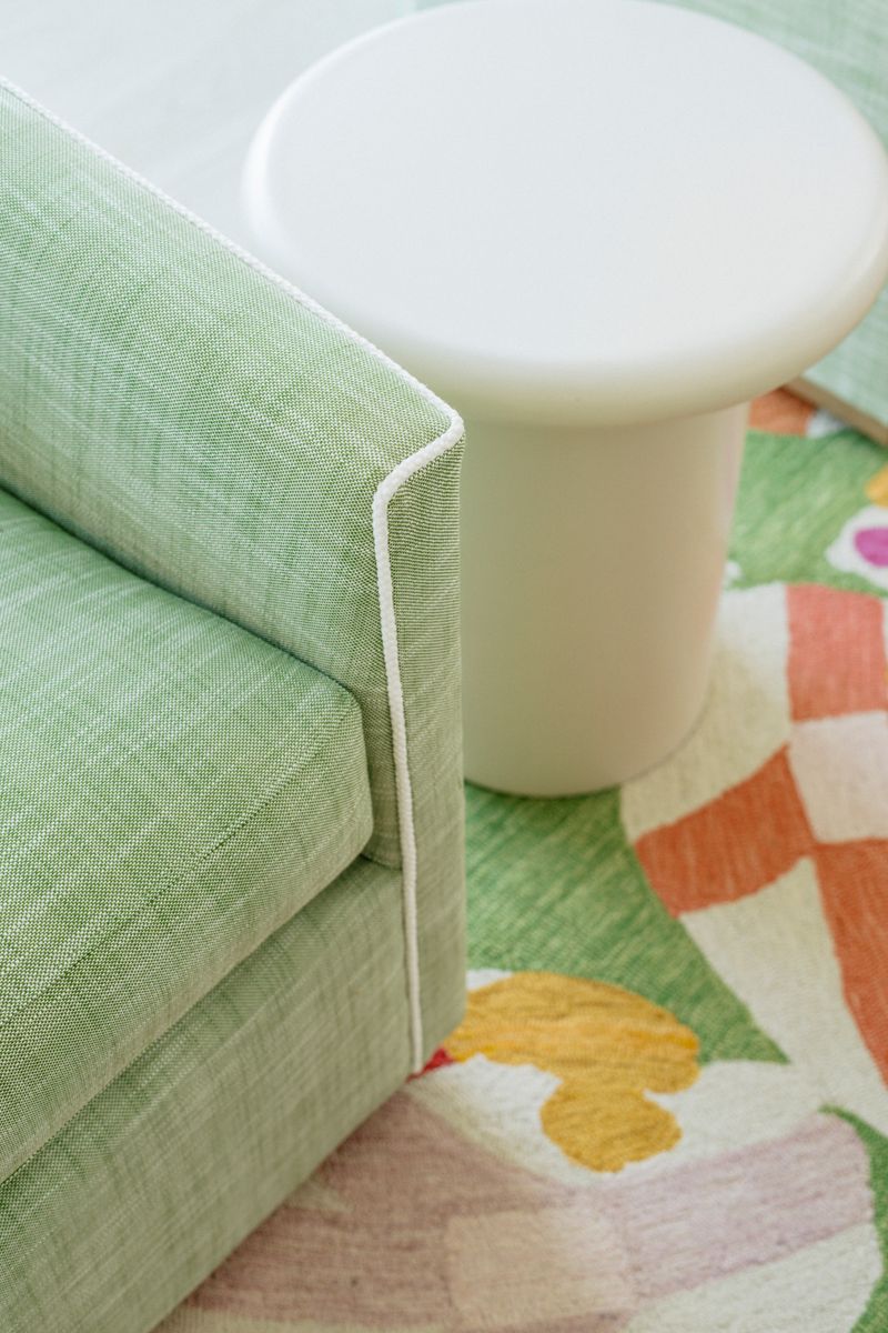 A living room in Panora with a green linen sofa trimmed in white piping beside a floral rug.