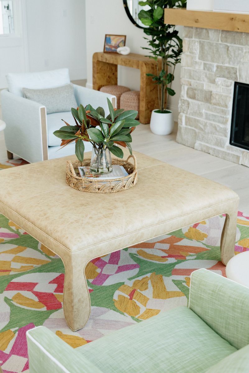 A living room in Panora with a cream leather ottoman on a colorful abstract rug, beside a limestone fireplace.