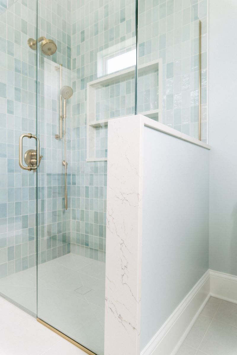 A primary bath shower in Panora tiled in sea glass blue with brushed brass fixtures and a marble half wall.