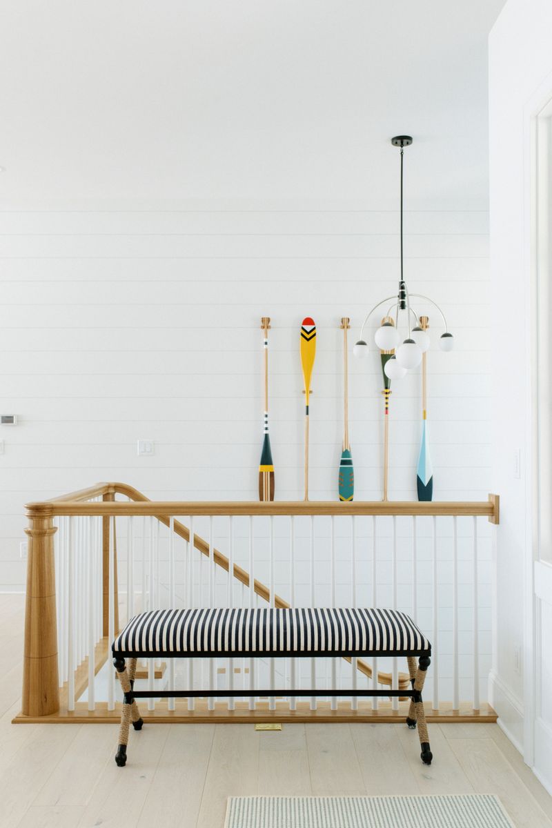 A stair landing in Panora with a black and white striped bench under painted wood oars on white shiplap.