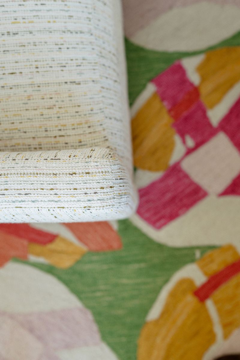 A cream flecked textured upholstery fabric in Panora, paired with a pink and green geometric patterned rug.